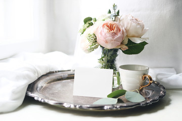 Spring, summer still life. Blank place card mockup, cup of coffee on old silver tray near window. Vintage feminine styled photo. Floral composition of pink English roses, Ranunculus and eucalyptus.