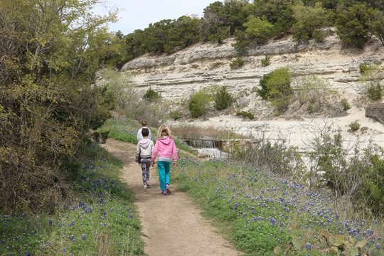 Three children hiking on a nature trail amidst bluebonnets, trees, limestone and a waterfall