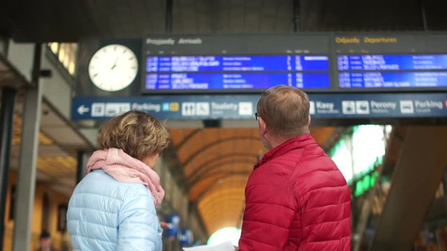 A middle-aged man and woman are waiting for a train on a train platform. Look at the schedule board up