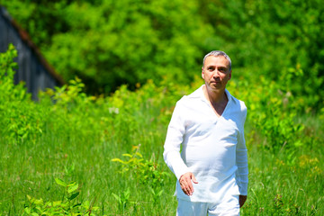 Naklejka na ściany i meble A man in white clothes walking in a field and touching flowers, surrounded by green.