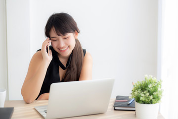 Beautiful young freelance asian woman smiling working on laptop computer and talking mobile smart phone at desk office with professional, girl using notebook, business and lifestyle concept.