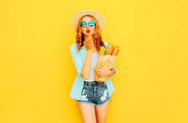 portrait beautiful woman sending sweet air kiss holding paper bag with long white bread baguette, wearing straw hat, shorts on colorful yellow background
