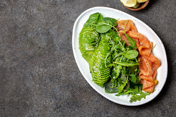Low carbs salad. Spinach, rucola salad with avocado and salmon. Black concrete background, white plate, top view