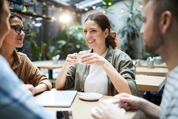 Happy excited young friends sitting at table in modern cafe and laughing while chatting and drinking coffee 