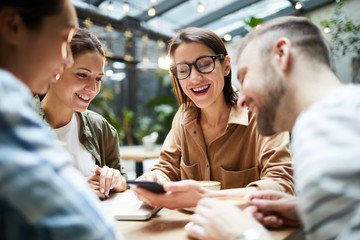 Positive excited young friends sitting at table and watching video on smartphone together while sharing news during gathering in cafe
