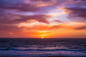 Fototapeta na wymiar Sun disappears beneath horizon over ocean after spectacular sunset with vivid sky. Wide angle scene at dusk.