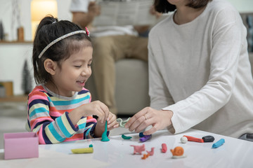 Mom with her daughter playing with a colorful toys modeling clay on the floor, at background daddy is sitting in a sofa in the living room, Happy asian family at home
