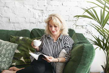 Indoor shot of fashionable attractive senior blonde female in striped shirt relaxing on comfortable...