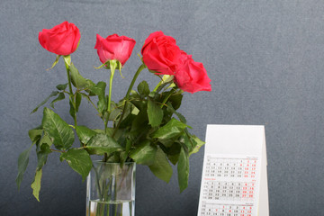 A bouquet of scarlet roses in a glass vase. Next is a calendar. On a gray background.