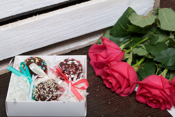 Cake pops are beautifully packed in a gift box. Nearby there is a cover with a transparent window and a bouquet of scarlet roses. On the background of wooden boards
