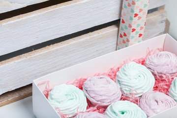 Homemade marshmallows of different colors are beautifully packed in a gift box. Nearby is the lid of the box with a transparent window. On the background of wooden boards.