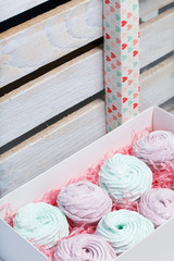 Homemade marshmallows of different colors are beautifully packed in a gift box. Nearby is the lid of the box with a transparent window. On the background of wooden boards.