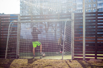 A boy teenager in shorts stands at the gate at the first spring sunshine after winter. The boys play in the courtyard on a specially equipped football field on the background of apartment buildings.