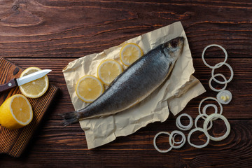 Fototapeta na wymiar Herring lies on paper with lemon slices, onion rings, parsley, dill. Salted herring lies on a brown wooden table. View from above.