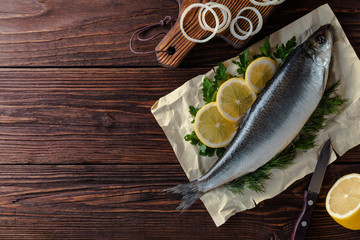 Herring lies on paper with lemon slices, onion rings, parsley, dill. Salted herring lies on a brown wooden table. View from above.