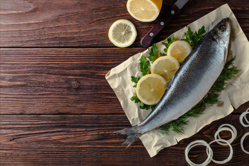 Herring lies on paper with lemon slices, onion rings, parsley, dill. Salted herring lies on a brown wooden table. View from above.