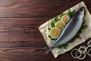 Herring lies on paper with lemon slices, onion rings, parsley, dill. Salted herring lies on a brown wooden table. View from above.