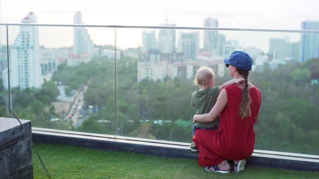 Son touches his mom for braid while she enjoys the view of a city and the beautiful sunset from the viewpoint.