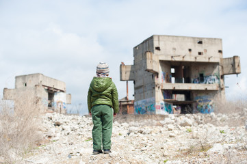 one little lonely child in green jacket standing on ruins of destroyed buildings in war zone