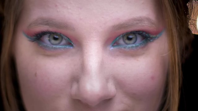 Eye-portrait of blonde model with colorful make-up joyfully ogling the camera on blurred lights background.