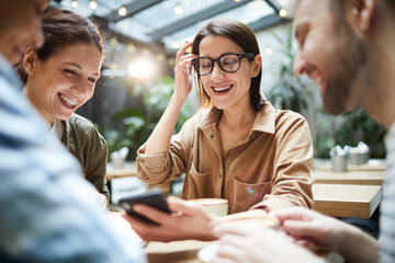 Cheerful attractive smart young woman in glasses sitting at table and showing online information while sharing idea for project with colleagues