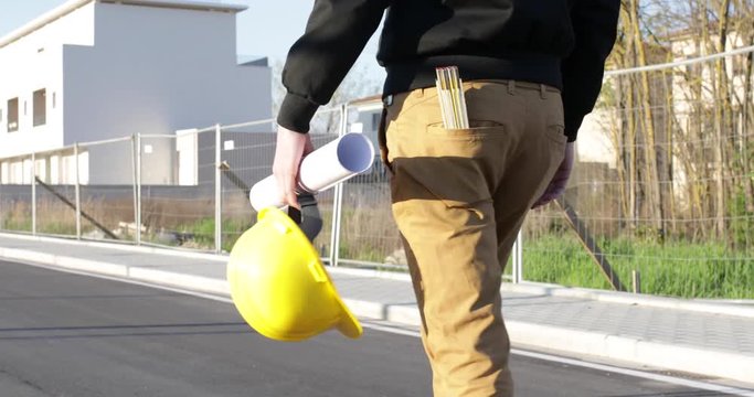 Giovane ingegnere cammina con il caschetto e il progetto sul foglio tenuti in mano. Vista posteriore delle gambe, pantaloni gialli, giacca nero e con metro nella tasca.