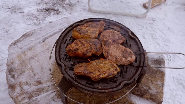 Steaks are roasted on camping portable grill, outdoor barbecue in winter, snow covered earth. Cast-iron burner with meat stands on wooden stump, hot coals visible through grate, top view stock video