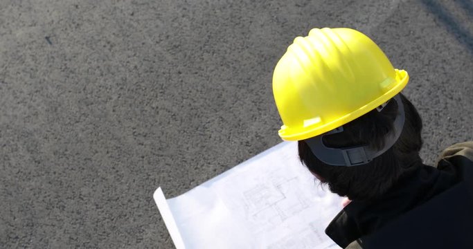 Ingegnere analizza il progetto su un foglio di carta bianco poggiato sulla strada mentre &egrave; in corso la revisione dei lavori. Vista della testa dall'alto con elmetto protettivo giallo.