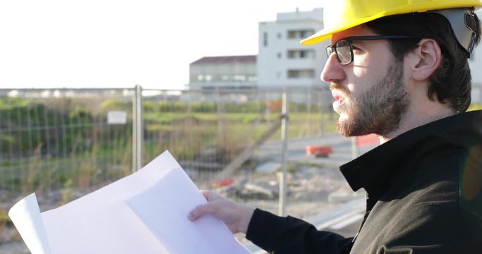Giovanissimo ingegnere edile con caschetto giallo, occhiali e barba sta osservando la realizzazione dei lavori sul suo progetto tenuto in mano.