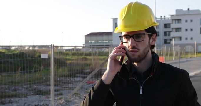 Ragazzo giovane architetto parla al telefono per affari di lavoro mentre &egrave; in cantiere con il casco protettivo giallo. Occhiali da vista, giacca colore nero e barba lunga. 