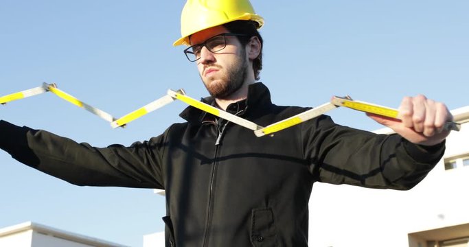 Giovane ragazzo ingegnere appena laureato con occhiali, barba, caschetto giallo, sta aprendo un metro di legno per misurare le costruzioni fatte dagli operai per valutarne la qualit&agrave; e la correttezza.