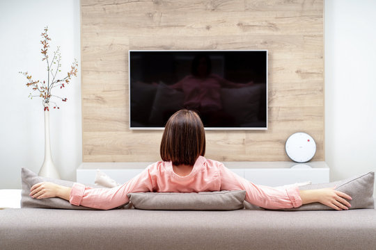 Back View Of Lonely Sad Depressive Beautiful Young Woman In Luxury Apartments, Sitting On The Sofa And Watching TV.