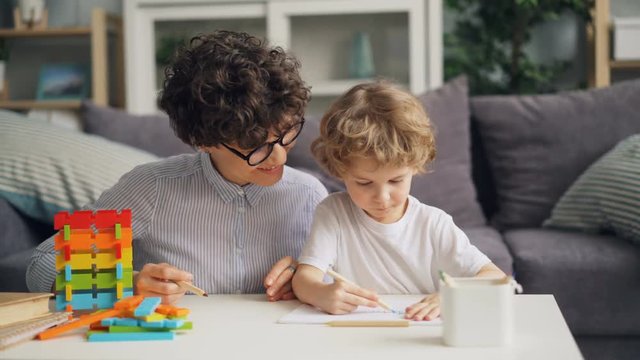 Creative little boy is drawing picture with pencils while cheerful woman is talking and smiling proud of her child. Family, creativity and leisure time concept.