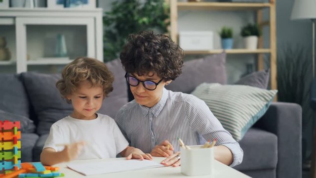 Smiling child cute boy is drawing with pencils while loving mother is talking and helping him in light cozy room. Hobby, childhood and creative people concept.