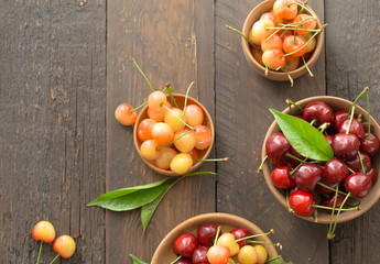 White cup with ripe berries of yellow and red sweet cherries and several fruit with green leaf of cherry tree on wooden background