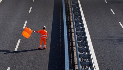 A highway worker with a high visibility work suit waves the orange flag to slow down traffic before...