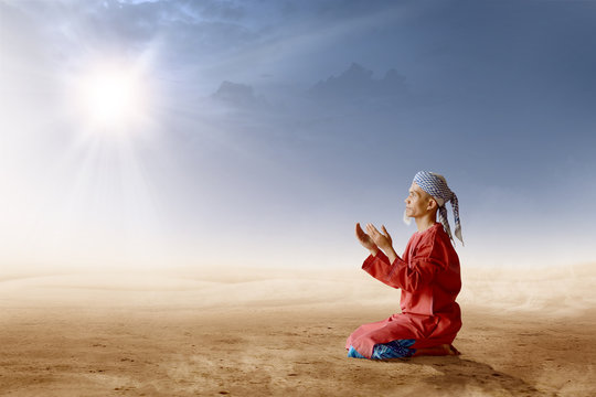 Asian Muslim Man With Turban In His Head Kneeling And Praying With Raised Hands On Desert