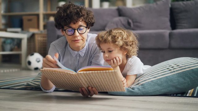 Happy family young woman and little boy are reading book and talking lying on blanket at home spending time together. Leisure and carefree childhood concept.