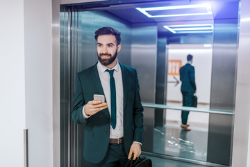 Smiling Caucasian businessman in formal wear holding briefcase and smart phone while getting out of elevator.