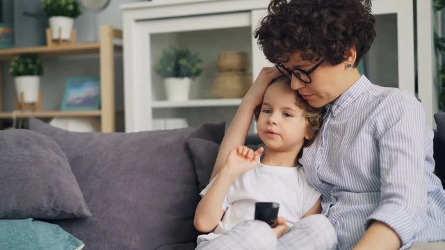Young woman loving mother is watching TV with cute little boy stroking and kissing him expressing love and tenderness. Parents, children and house concept.