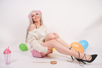 Obraz na płótnie Canvas cute blonde caucasian happy girl dressed in a milky color knitted sweater and funny shorts, she sits on the white floor alone with balloons and a glass of water 