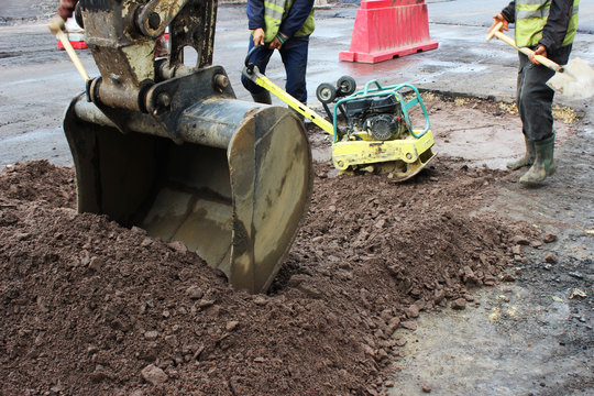 The foreman of the construction team works with a vibrating platform plate, tamping up the sand before the asphalting of the road using the patch method. reportage. foreground excavator bucket.
