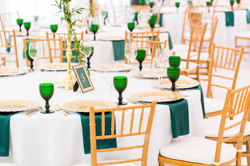 Interior of tent for wedding dinner, ready for guests. Served round banquet table. Golden dishes, green wine glasses and napkins. Catering concept.