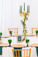Interior of tent for wedding dinner, ready for guests. Served round banquet table. Golden dishes, green wine glasses and napkins. Catering concept.