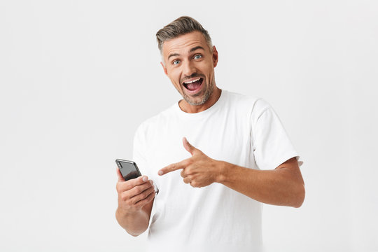 Image of optimistic man 30s wearing casual t-shirt rejoicing while using smartphone