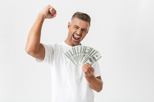 Image of unshaved man 30s wearing casual t-shirt celebrating while holding bunch of money banknotes