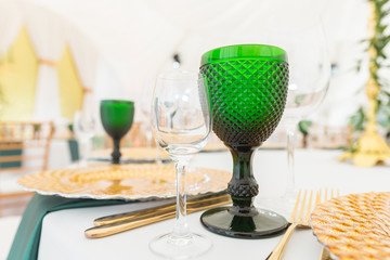 Golden dishes, green wine glasses and napkins. Catering concept. Interior of tent for wedding dinner, ready for guests. Served round banquet table.