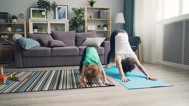 Young mother and little son are doing yoga together at home relaxing enjoying activity on mat. Proud mom is smiling looking at child with pride and love.