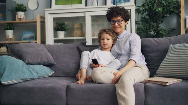 Happy child is watching TV with loving mother at home on sofa holding remote control embracing and laughing. Happiness, family and modern technology concept.