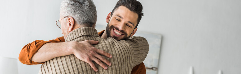 panoramic shot of happy bearded man smiling while hugging elder father at home